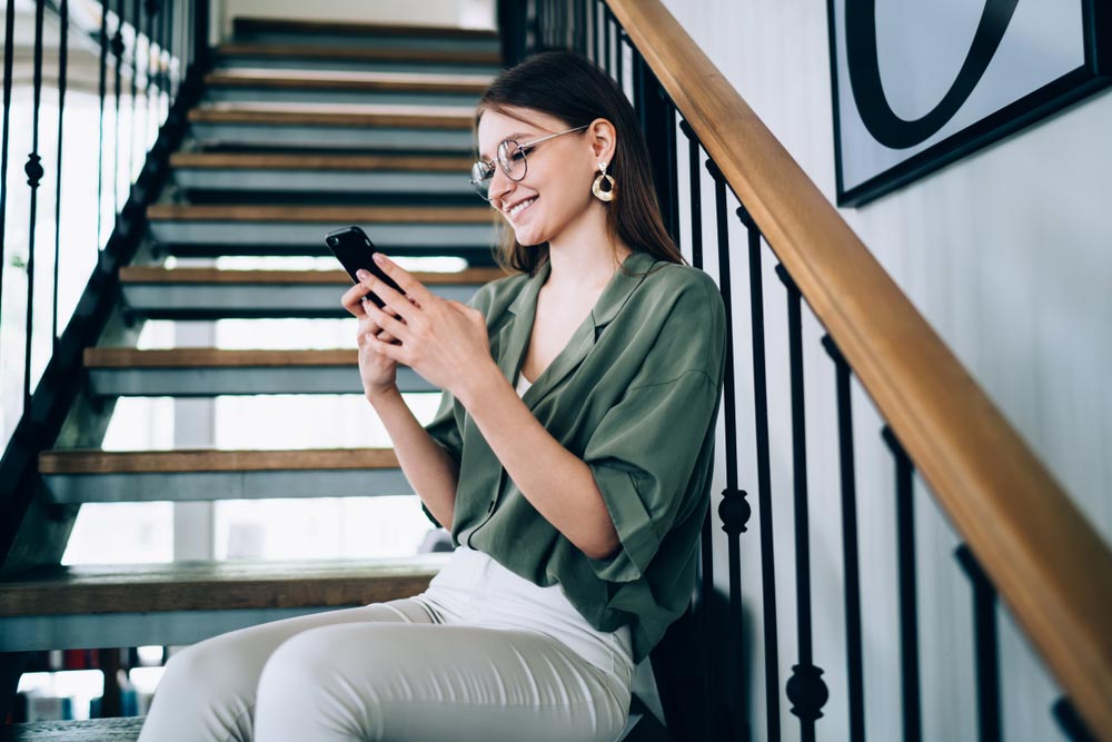 Happy content woman in comfy blouse and spectacles resting on decorative stairs and using smartphone in room with stylish interior