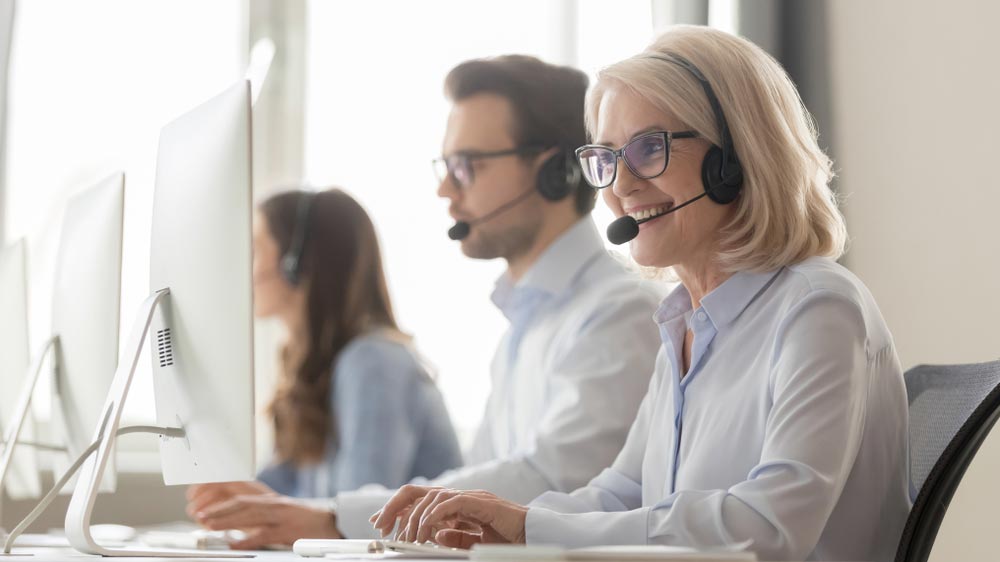 Smiling old female call center agent operator telemarketer in headset talking consulting online client at workplace